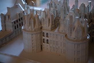 A detailed architectural model of a grand, intricate building featuring towers, domes, and ornate details. The model is crafted in a monochromatic color scheme, giving it an elegant and timeless appearance. The structure showcases impressive craftsmanship with its numerous windows, arches, and decorative elements.