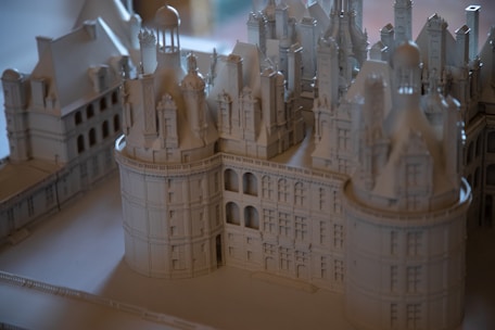 A detailed architectural model of a grand, intricate building featuring towers, domes, and ornate details. The model is crafted in a monochromatic color scheme, giving it an elegant and timeless appearance. The structure showcases impressive craftsmanship with its numerous windows, arches, and decorative elements.
