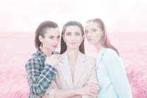 Three women stand closely together in a field of tall grass with pinkish hues. Each of them is wearing a distinct blazer in patterns and shades ranging from checkered to solid. They have calm and serene expressions, and their hairstyles are sleek and styled back. The background is softly blurred, enhancing the dreamy and ethereal quality of the scene.