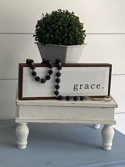 A small white table with turned legs holds a decorative sign with the word 'grace' and a dark bead garland draped across it. Above the sign, there is a potted plant with lush green foliage in a white ceramic planter.