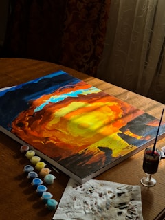 A canvas rests on a wooden table, displaying a vibrant painting of a swirling, fiery sunset with intense oranges and yellows. Surrounding the canvas are small containers of paint arranged in a line. Sunlight streams through a window, partially illuminating the scene, creating a warm atmosphere. A paintbrush is placed in a glass filled with water, while a stained paper towel lies nearby.