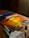 A canvas rests on a wooden table, displaying a vibrant painting of a swirling, fiery sunset with intense oranges and yellows. Surrounding the canvas are small containers of paint arranged in a line. Sunlight streams through a window, partially illuminating the scene, creating a warm atmosphere. A paintbrush is placed in a glass filled with water, while a stained paper towel lies nearby.