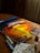 A canvas rests on a wooden table, displaying a vibrant painting of a swirling, fiery sunset with intense oranges and yellows. Surrounding the canvas are small containers of paint arranged in a line. Sunlight streams through a window, partially illuminating the scene, creating a warm atmosphere. A paintbrush is placed in a glass filled with water, while a stained paper towel lies nearby.