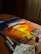 A canvas rests on a wooden table, displaying a vibrant painting of a swirling, fiery sunset with intense oranges and yellows. Surrounding the canvas are small containers of paint arranged in a line. Sunlight streams through a window, partially illuminating the scene, creating a warm atmosphere. A paintbrush is placed in a glass filled with water, while a stained paper towel lies nearby.