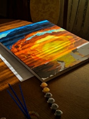 A partially completed canvas painting lies on a wooden table, capturing a vibrant scene with swirling, fiery colors resembling a sunset or cosmic vortex. Sunlight casts a warm glow across the artwork, enhancing the deep blues, oranges, and yellows. Nearby, small containers of paint and brushes are neatly arranged, indicating ongoing creative work.