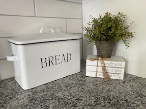 A bamboo bread box sitting on a kitchen counter beside freshly baked bread and herbs.