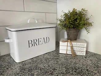 A bamboo bread box sitting on a kitchen counter beside freshly baked bread and herbs.