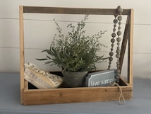 A rustic wooden frame holds a green potted plant with delicate leaves. Beside the plant, a small blue sign with the words 'live simply' is placed inside the frame. A stack of books wrapped with a decorative ribbon lies on the left, while a string of wooden beads hangs from the right corner.