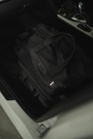 A black duffel bag is placed on the black leather seat of a car. The bag has dual handles and a small rectangular logo with red, white, and blue stripes. The car interior appears modern with a visible gear shift and center console.