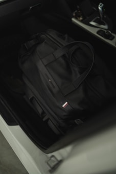 A black duffel bag is placed on the black leather seat of a car. The bag has dual handles and a small rectangular logo with red, white, and blue stripes. The car interior appears modern with a visible gear shift and center console.
