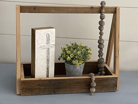A wooden display crate containing a tall, rectangular block with a carved cross, a small metal pot with green and yellow foliage, and a strand of large, round wooden beads hanging over the side.