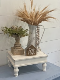 A rustic vignette featuring a white pitcher filled with dried wheat stalks and a wooden bird perched on a decorative stand with green foliage. A small wooden sign with the text 'Our Nest' is placed on a white, vintage-style stand. The overall aesthetic is cozy and farmhouse-inspired.