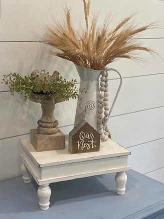 A rustic vignette featuring a white pitcher filled with dried wheat stalks and a wooden bird perched on a decorative stand with green foliage. A small wooden sign with the text 'Our Nest' is placed on a white, vintage-style stand. The overall aesthetic is cozy and farmhouse-inspired.