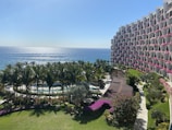 A resort-like setting with an expansive view of the ocean under a clear blue sky. Palm trees line a pool area, and a large building with pink flowers on the balconies curves around on the right.