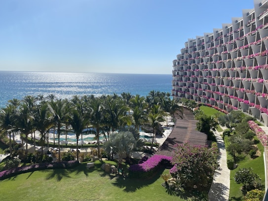 A resort-like setting with an expansive view of the ocean under a clear blue sky. Palm trees line a pool area, and a large building with pink flowers on the balconies curves around on the right.