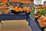 A table covered with a blue cloth displays a variety of fresh fruits and vegetables. On the left, there are baskets filled with brown paper bags and a metal bowl containing oranges. The middle of the table features bowls with apples, bananas, and informational pamphlets. To the right, packaged salad greens and leafy vegetables like beets are arranged in bowls. People are visible in the background, suggesting a market or community event setting.