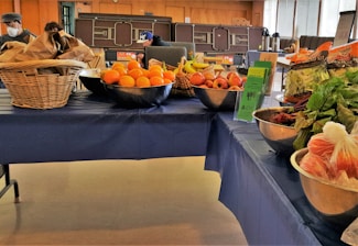 A table covered with a blue cloth displays a variety of fresh fruits and vegetables. On the left, there are baskets filled with brown paper bags and a metal bowl containing oranges. The middle of the table features bowls with apples, bananas, and informational pamphlets. To the right, packaged salad greens and leafy vegetables like beets are arranged in bowls. People are visible in the background, suggesting a market or community event setting.