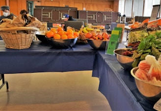 A table covered with a blue cloth displays a variety of fresh fruits and vegetables. On the left, there are baskets filled with brown paper bags and a metal bowl containing oranges. The middle of the table features bowls with apples, bananas, and informational pamphlets. To the right, packaged salad greens and leafy vegetables like beets are arranged in bowls. People are visible in the background, suggesting a market or community event setting.
