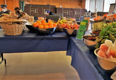 A table covered with a blue cloth displays a variety of fresh fruits and vegetables. On the left, there are baskets filled with brown paper bags and a metal bowl containing oranges. The middle of the table features bowls with apples, bananas, and informational pamphlets. To the right, packaged salad greens and leafy vegetables like beets are arranged in bowls. People are visible in the background, suggesting a market or community event setting.