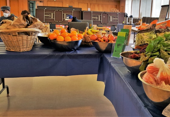 A table covered with a blue cloth displays a variety of fresh fruits and vegetables. On the left, there are baskets filled with brown paper bags and a metal bowl containing oranges. The middle of the table features bowls with apples, bananas, and informational pamphlets. To the right, packaged salad greens and leafy vegetables like beets are arranged in bowls. People are visible in the background, suggesting a market or community event setting.