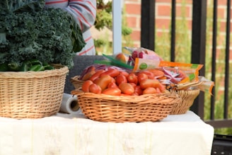 A wicker basket filled with fresh tomatoes and packaged vegetables is placed on a table. Another basket contains leafy greens, possibly kale and green peppers. The setting appears to be outdoors, and a person wearing a striped shirt is partially visible behind the produce.