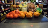 A variety of fruits and vegetables placed on a checkout conveyor belt, including oranges in a mesh bag, pears, lemons, and packaged vegetables.