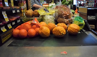 A variety of fruits and vegetables placed on a checkout conveyor belt, including oranges in a mesh bag, pears, lemons, and packaged vegetables.