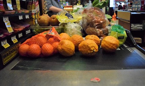 A variety of fruits and vegetables placed on a checkout conveyor belt, including oranges in a mesh bag, pears, lemons, and packaged vegetables.