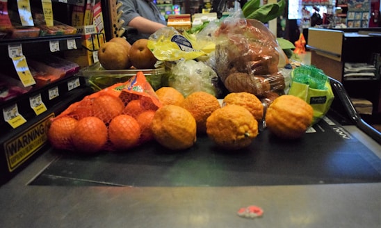 A variety of fruits and vegetables placed on a checkout conveyor belt, including oranges in a mesh bag, pears, lemons, and packaged vegetables.