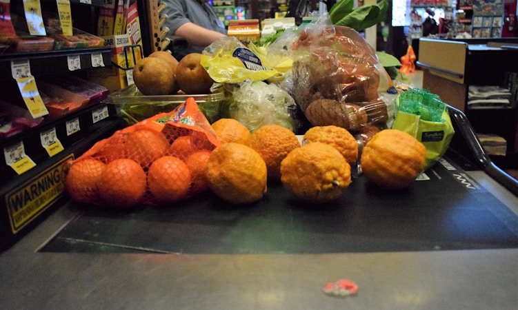 A variety of fruits and vegetables placed on a checkout conveyor belt, including oranges in a mesh bag, pears, lemons, and packaged vegetables.