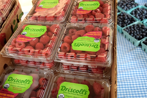 Several plastic containers filled with Driscoll's organic raspberries are stacked together. Each container is labeled with green stickers indicating the brand and organic certification. To the side, there are smaller cardboard containers filled with blueberries. The background features a blue and white checkered cloth.