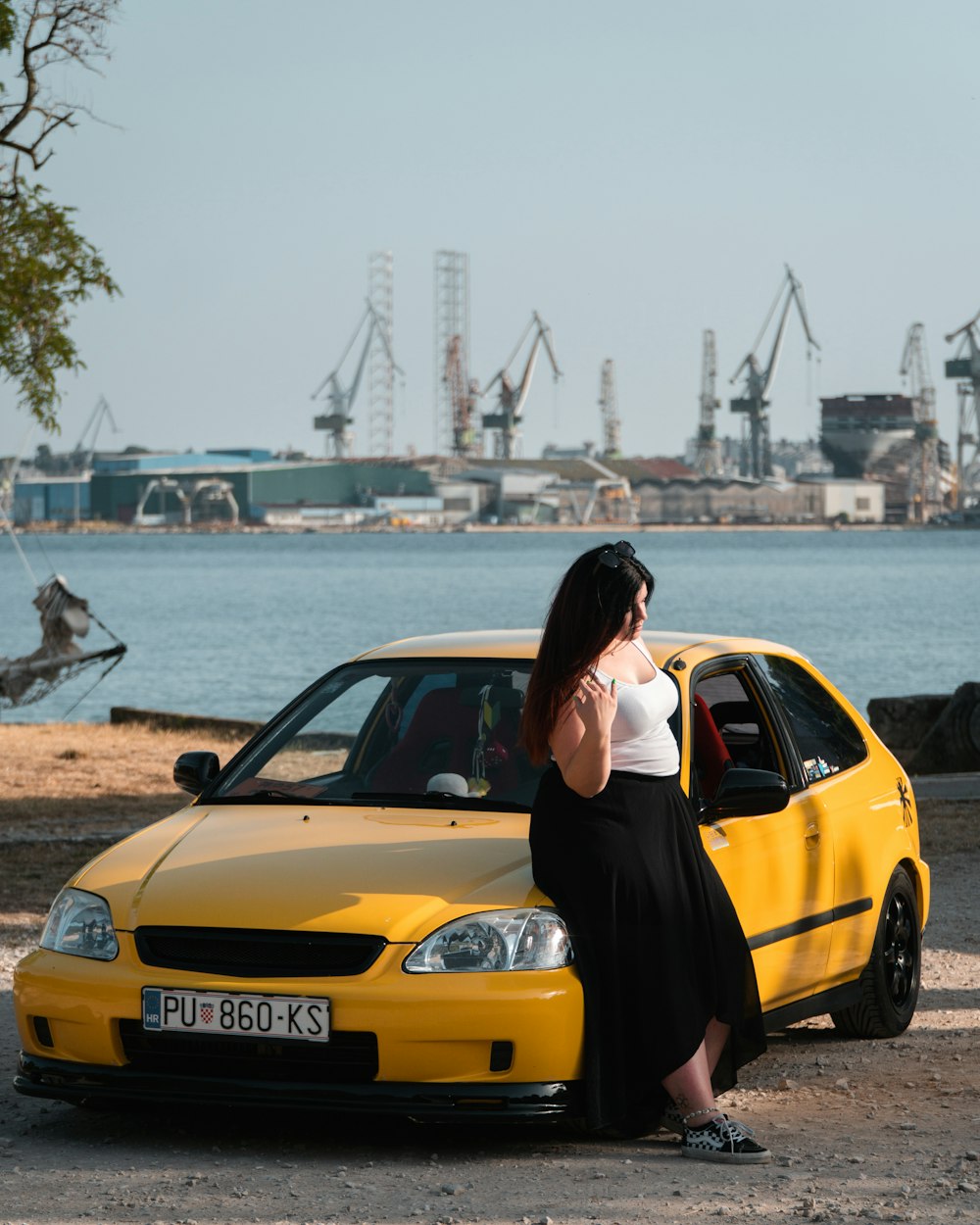 a woman leaning on a yellow car in front of a body of water