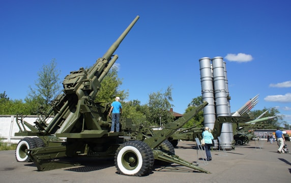 A large military artillery piece is displayed outdoors, surrounded by trees and a clear blue sky. Several cylindrical missile launchers are visible in the background along with a few people observing them.