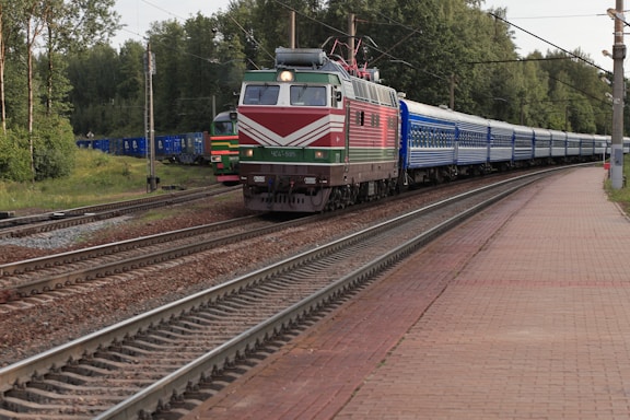 A train with a red and green locomotive pulling a long line of blue passenger cars runs along multiple sets of train tracks. The tracks are bordered by trees and green foliage, and a station platform is visible on the right side with a brick surface.
