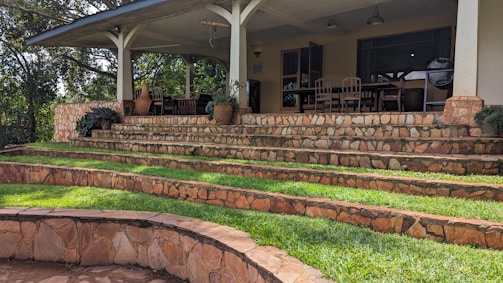 A series of stone steps with grass-covered terraces lead up to a patio area. The patio is partially covered by a roof supported by pillars and features wooden chairs around tables. Clay pots with plants are placed along the steps, adding to the landscaped design. Trees and greenery surround the setting, creating a peaceful outdoor space.