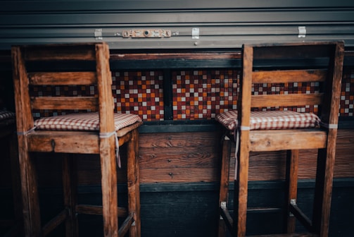 A pair of wooden bar stools with checked cushions are positioned at a high table. The background features a colorful mosaic tile design with a mix of red, orange, and white tiles. The setting appears to be rustic and cozy.
