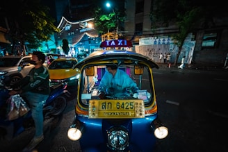 A nighttime street scene featuring a brightly lit tuk-tuk taxi with a driver inside. Neon lights illuminate the surroundings, including a sign for Chinatown. A motorcyclist wearing a helmet and mask rides by, and several cars are visible in the background. The atmosphere has a bustling urban vibe.