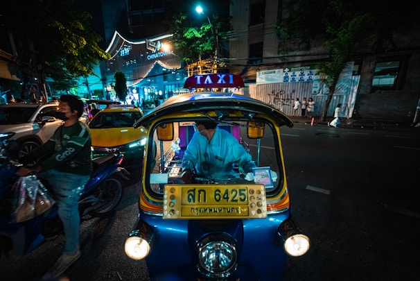 A nighttime street scene featuring a brightly lit tuk-tuk taxi with a driver inside. Neon lights illuminate the surroundings, including a sign for Chinatown. A motorcyclist wearing a helmet and mask rides by, and several cars are visible in the background. The atmosphere has a bustling urban vibe.