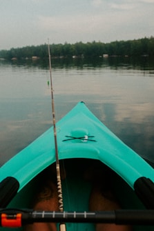 A turquoise kayak is floating on calm water with a fishing rod extending from it. In the background, a line of trees is visible along the shoreline under a cloudy sky.