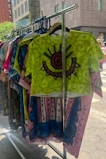 A colorful rack of T-shirts featuring abstract patterns and bold colors is displayed outdoors. The front shirt is bright green with an eye-like design in the center, surrounded by intricate patterns in red and black. Other shirts behind it display varying designs and vibrant colors, adding to the artistic vibe.