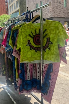 A colorful rack of T-shirts featuring abstract patterns and bold colors is displayed outdoors. The front shirt is bright green with an eye-like design in the center, surrounded by intricate patterns in red and black. Other shirts behind it display varying designs and vibrant colors, adding to the artistic vibe.