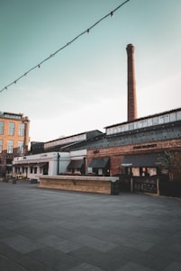A quiet, open plaza features a large industrial brick chimney rising into a clear sky. Surrounding buildings are constructed with a mix of brick and modern materials, appearing to be shops or restaurants, with signs and awnings. String lights are hanging across the scene, suggesting a lively atmosphere during night or events.