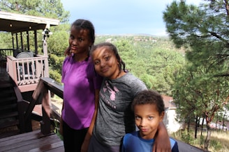 Three children stand on a wooden deck surrounded by a lush green forest. The deck has a railing and a small seating area with a table and chairs on the left. The children are closely huddled together with their arms around each other, smiling at the camera. In the background is a scenic view of trees and hills under a clear sky.