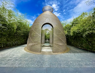 A large, sculptural dome structure made of horizontal wooden slats stands in the center of a stone path. It features a doorway leading to an interior with a circular opening at the top and a small stone bench or platform inside. Lush green bamboo plants surround the structure on both sides, under a bright blue sky with scattered white clouds.