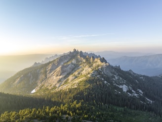 A panoramic view of the sun rising over the rugged peaks of the Alpes-Maritimes, with lush green forests in the foreground.