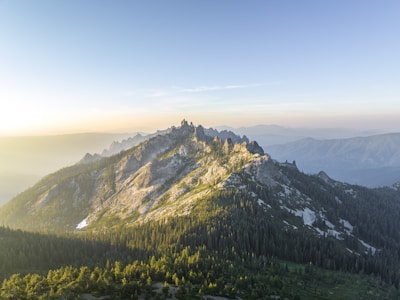 A panoramic view of the sun rising over the rugged peaks of the Alpes-Maritimes, with lush green forests in the foreground.
