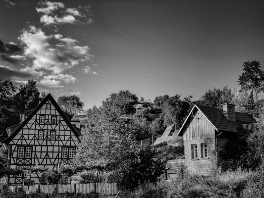 A black and white image of a countryside scene featuring two traditional houses surrounded by trees and bushes. The larger house displays a classic timber-framed architecture with patterned walls and multiple windows, while the smaller structure appears to be made of wood and stone. Above, the sky is partly cloudy, adding a dramatic element to the scene with its distinct cloud formations.
