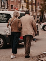 An elderly couple walking together on a city street, holding each other closely. They are wearing casual clothing and appear to be engaged in a leisurely stroll. Surrounding them are parked cars and urban buildings, with a soft, warm ambiance.