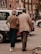 An elderly couple walking together on a city street, holding each other closely. They are wearing casual clothing and appear to be engaged in a leisurely stroll. Surrounding them are parked cars and urban buildings, with a soft, warm ambiance.