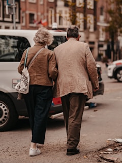 An elderly couple walking together on a city street, holding each other closely. They are wearing casual clothing and appear to be engaged in a leisurely stroll. Surrounding them are parked cars and urban buildings, with a soft, warm ambiance.